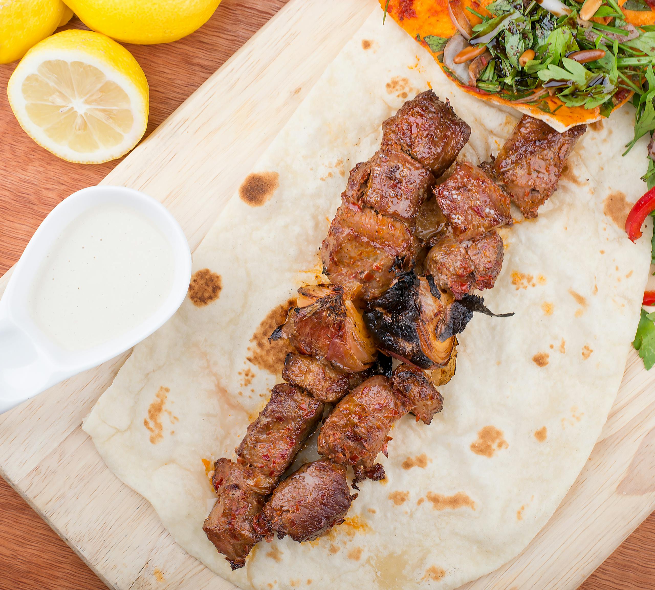 Close-up of grilled kebab on flatbread with fresh salad, lemon, and sauce.